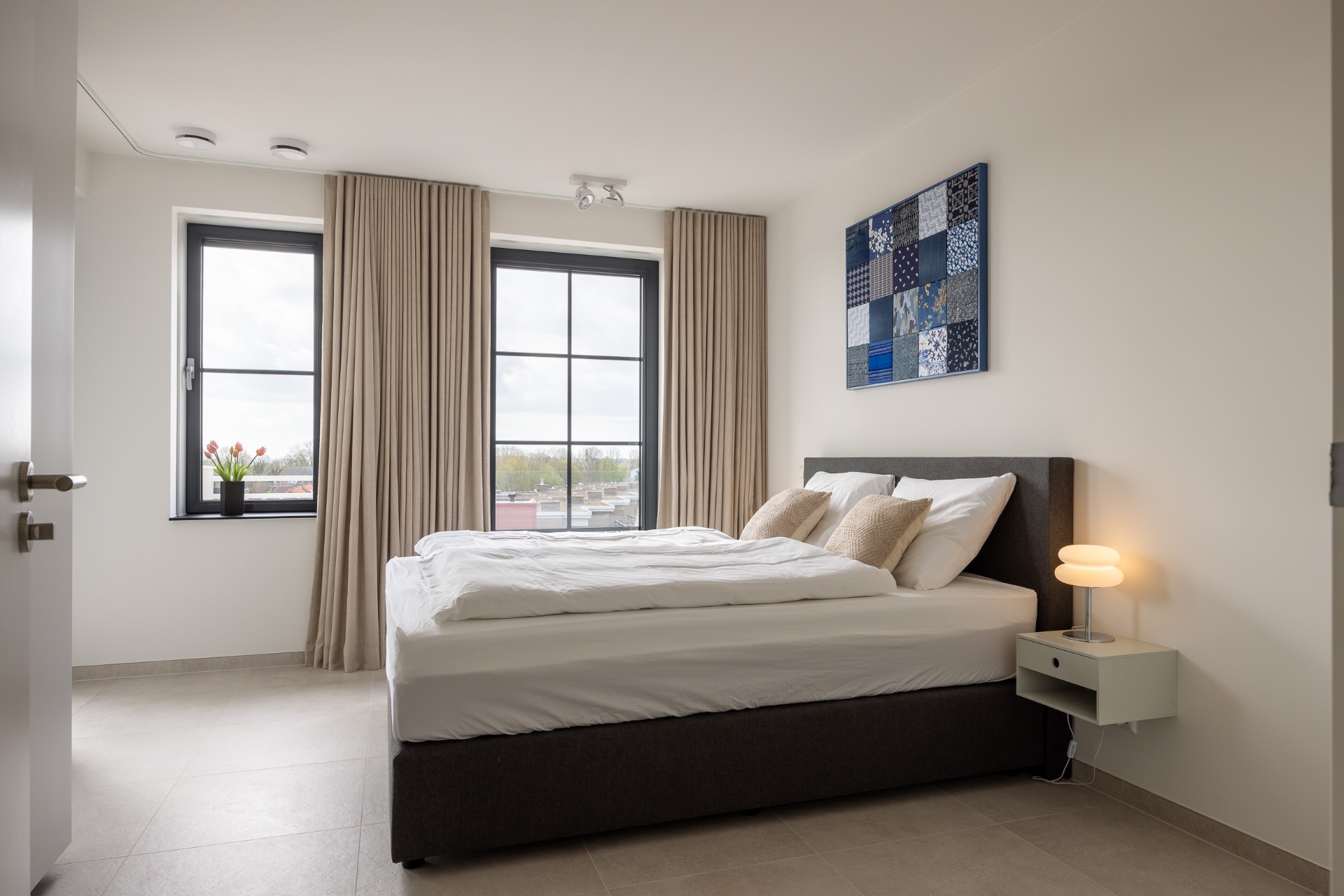 Lichte slaapkamer met tweepersoonsbed, beige gordijnen en blauw kunstwerk in vakantieappartement Cadzand-Bad